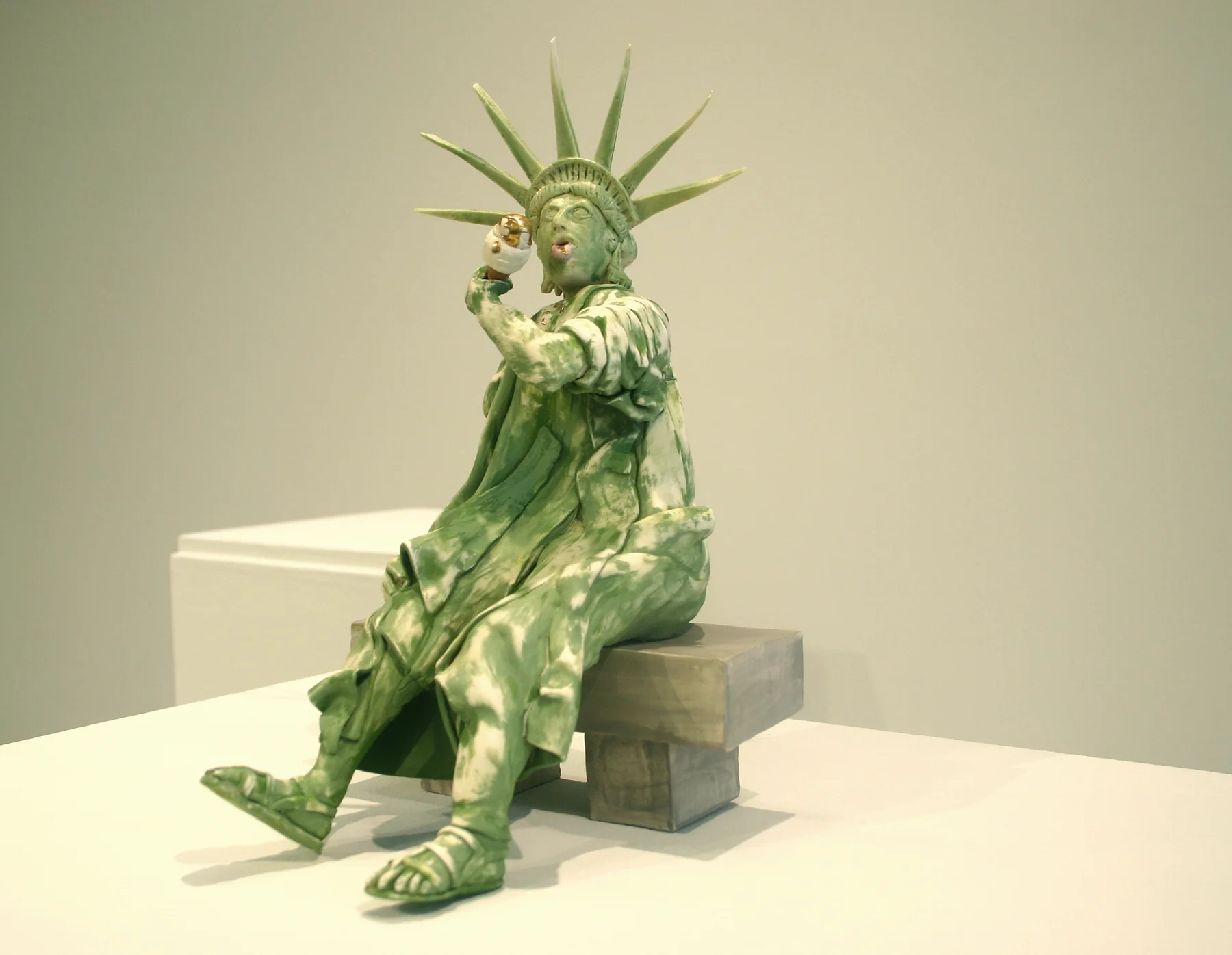 A seated ceramic figure of the Statue of Liberty glazed in weathered green, slumped forward on a plinth with her crown and torch.