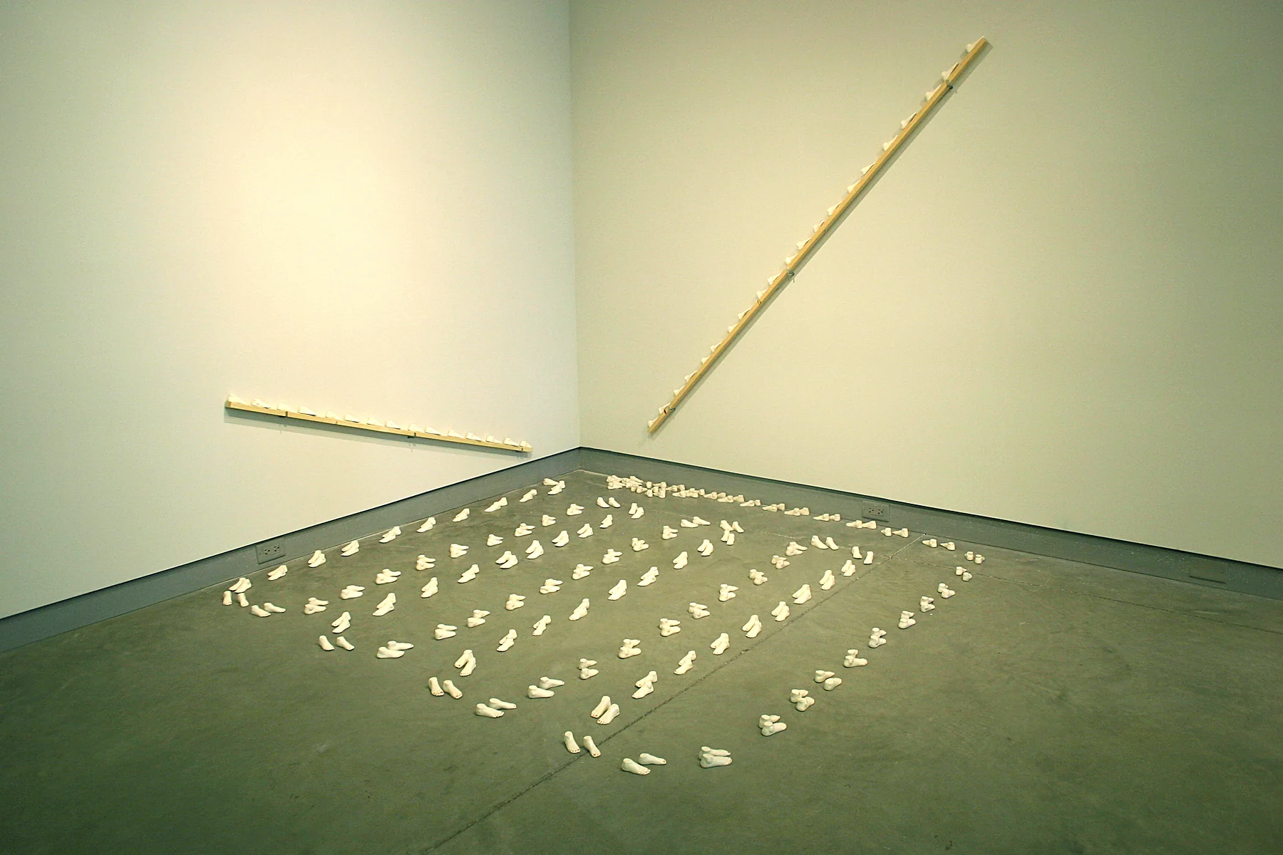 An installation view in a gallery corner: rows of small pale ceramic feet arranged in a grid across the floor, with a thin gold line running along the wall above.