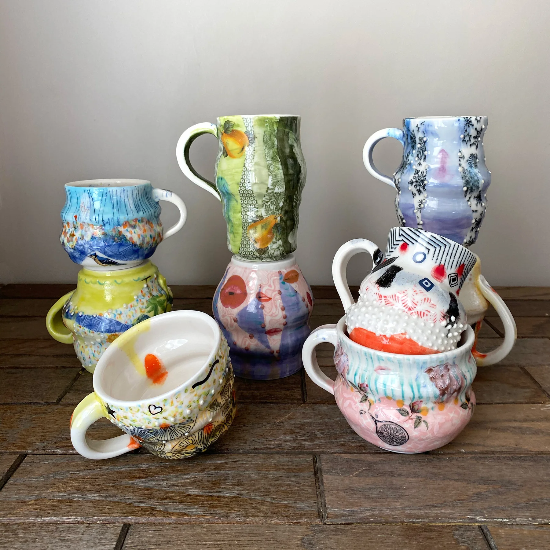 Seven hand-thrown porcelain mugs stacked in three columns, each with a different maximalist surface — birds, fruit, snowflakes, lips and raised white dots.